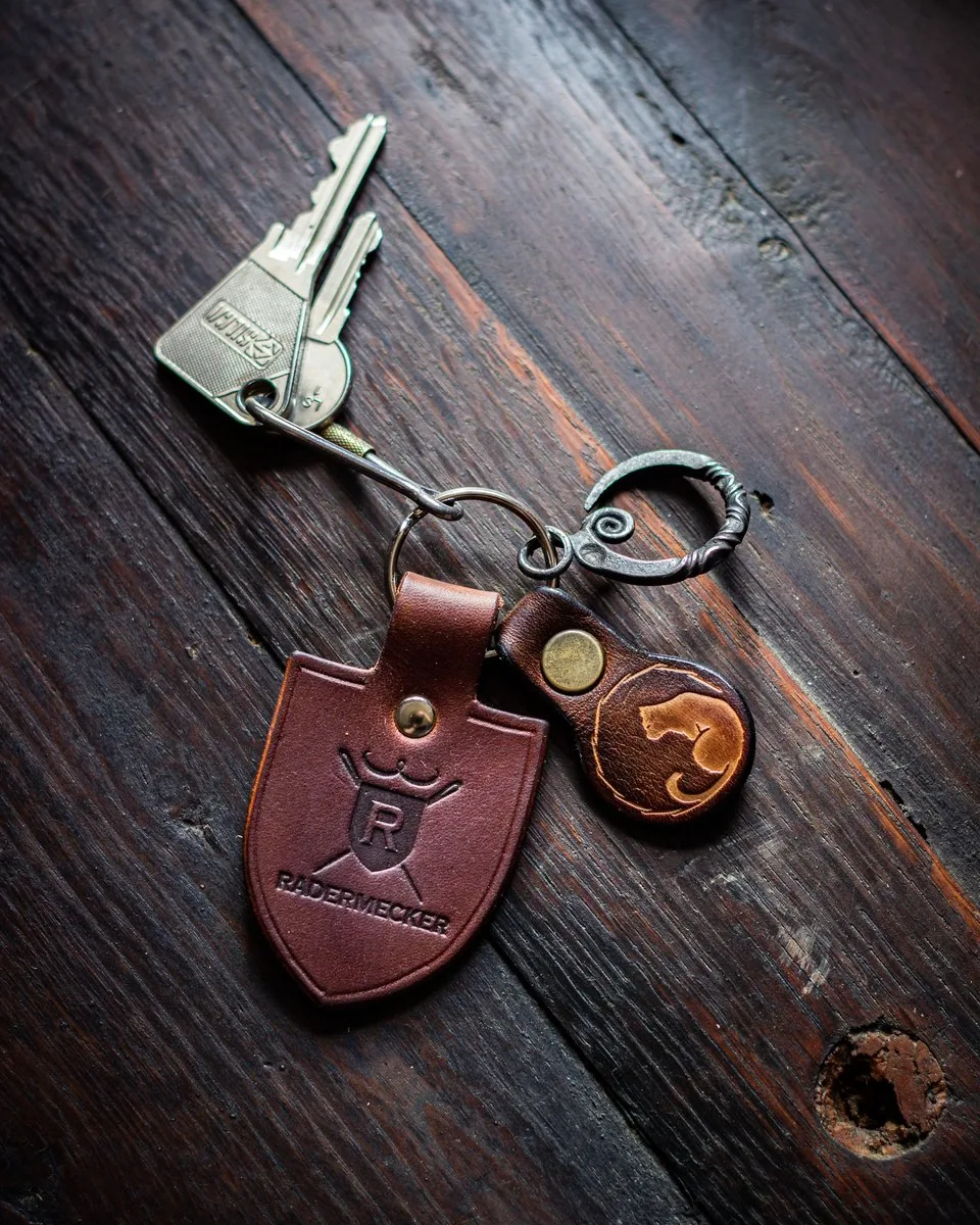 Deux porte-clés en cuir sur un trousseau, l’un en forme de blason marqué d’un monogramme, l’autre rond au chat de l’atelier, avec un anneau en fer forgé, sur un plancher sombre