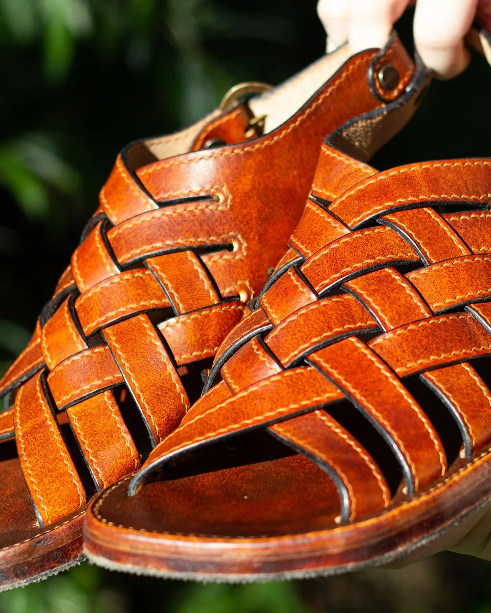 Paire de sandales en cuir fauve aux lanières tressées, semelles cousues, tenue à la main devant un feuillage
