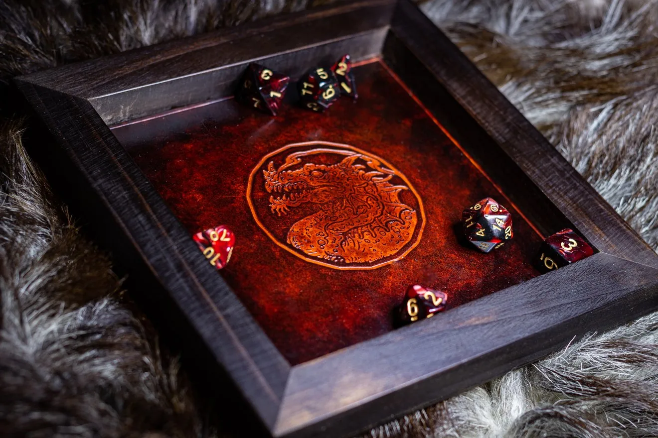 Plateau de dés en cuir teinté rouge, repoussé d’un dragon dans un médaillon, serti dans un cadre en bois sombre, avec des dés posés dessus