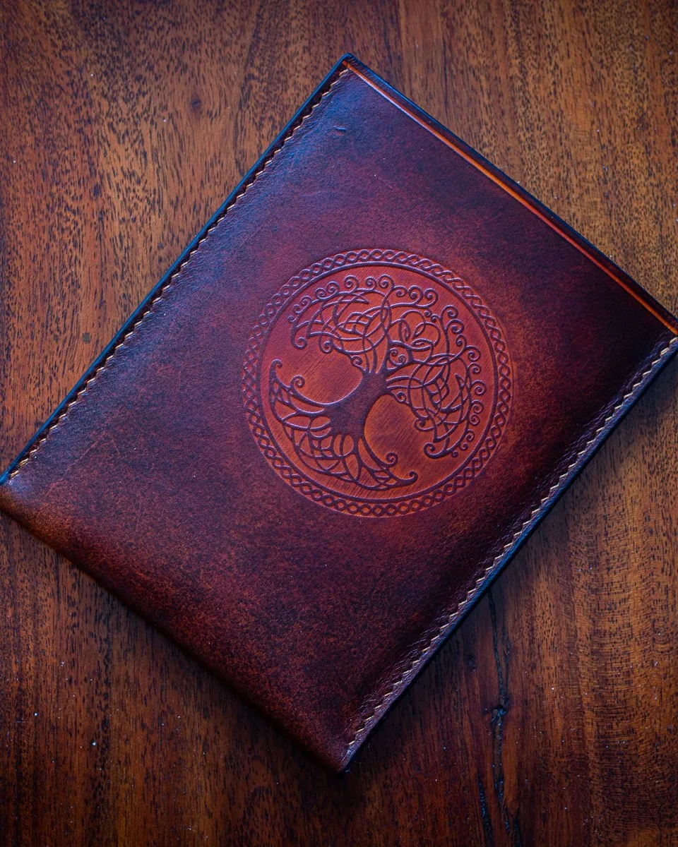 Pochette pour liseuse en cuir teinté bordeaux, repoussée d’un arbre de vie celtique dans un médaillon, posée sur une table en bois
