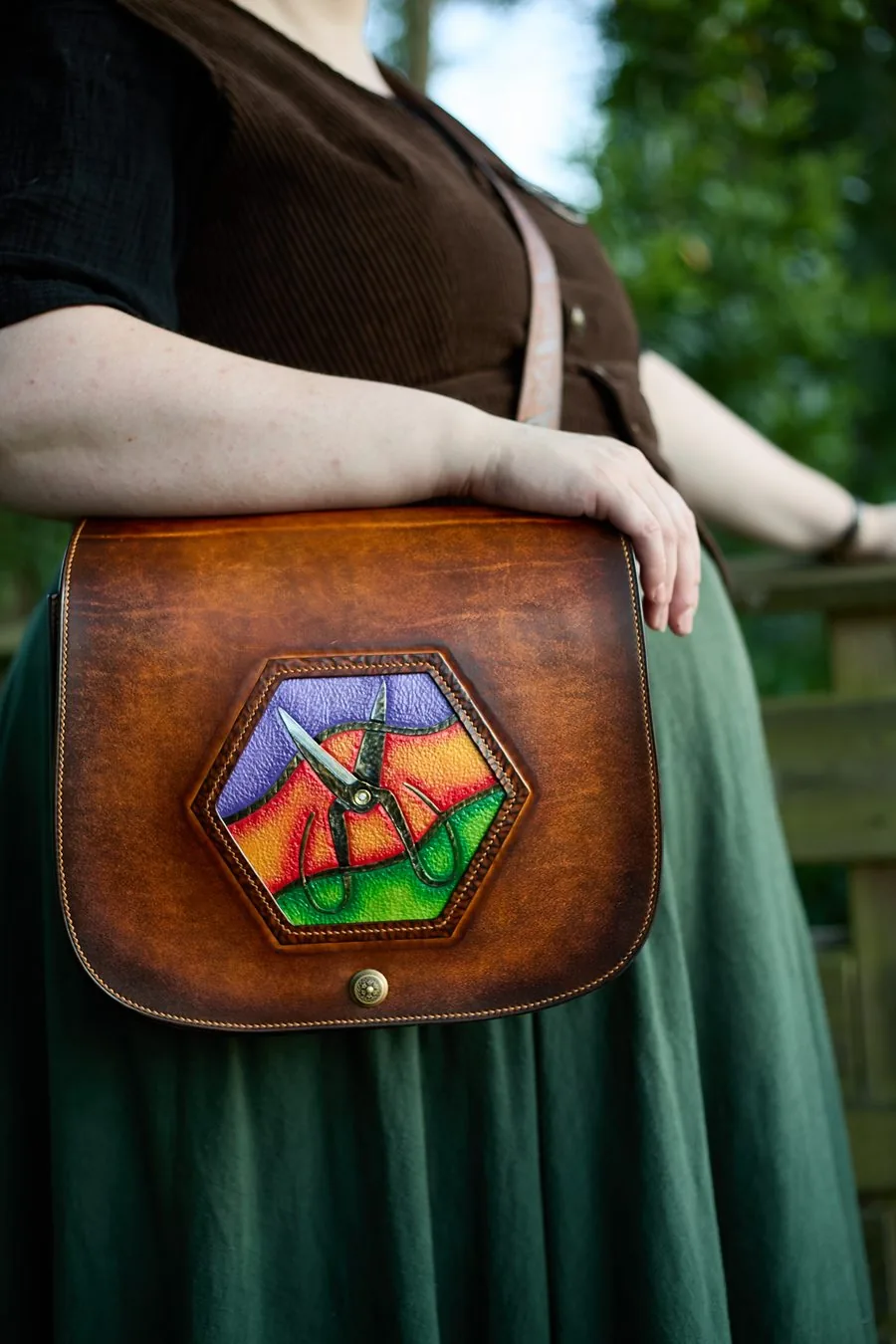 Sac à bandoulière en cuir brun porté à l'épaule, rabat orné d'un médaillon hexagonal peint de couleurs vives avec une paire de ciseaux en relief