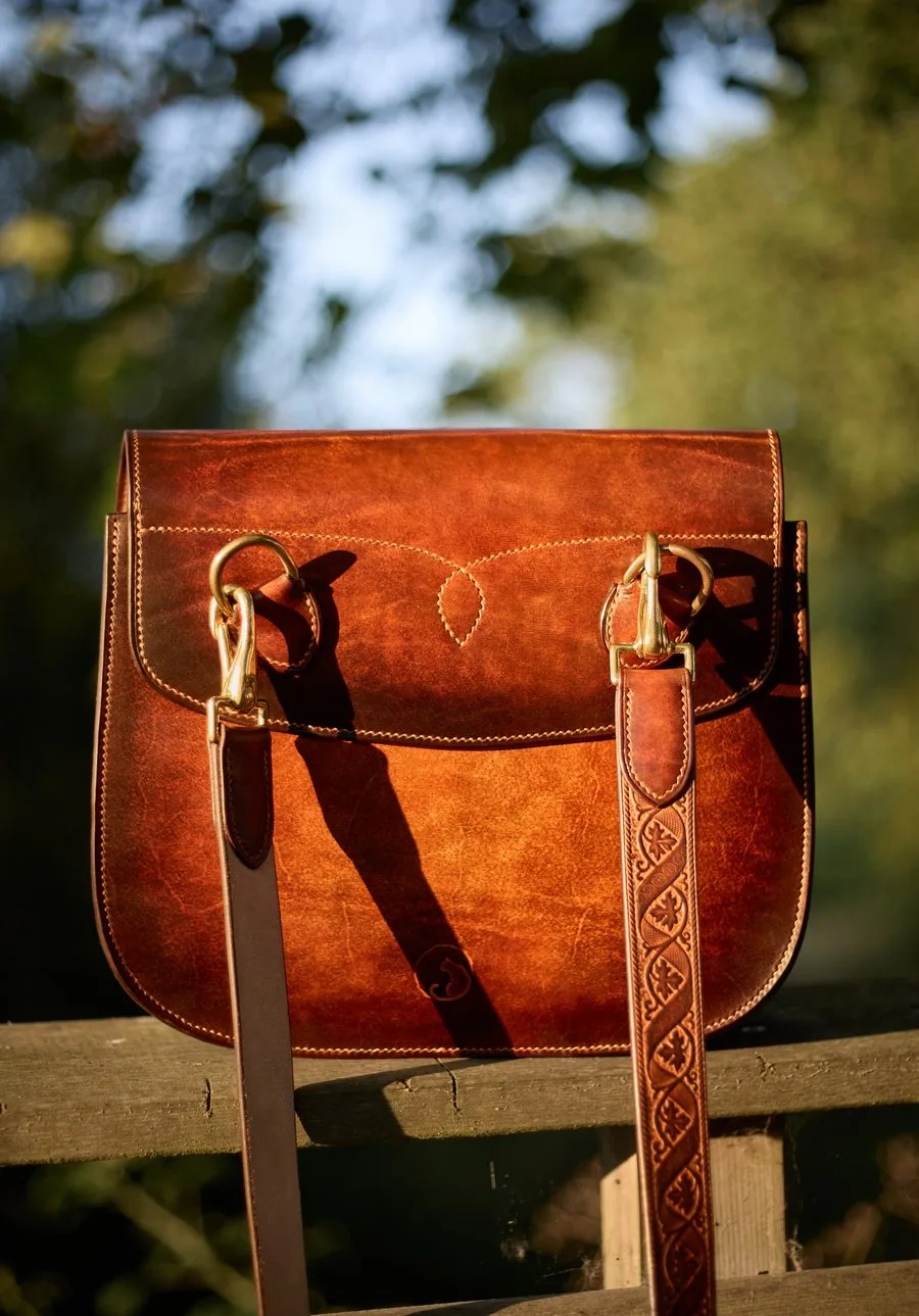 Sac en cuir fauve avec sa bandoulière ornée et ses mousquetons en laiton, posé sur une barrière